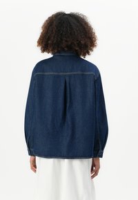 Persoon met krullend haar, gekleed in een donkerblauwe spijkerjacket over een witte rok, staat met de rug naar de camera tegen een effen achtergrond.
