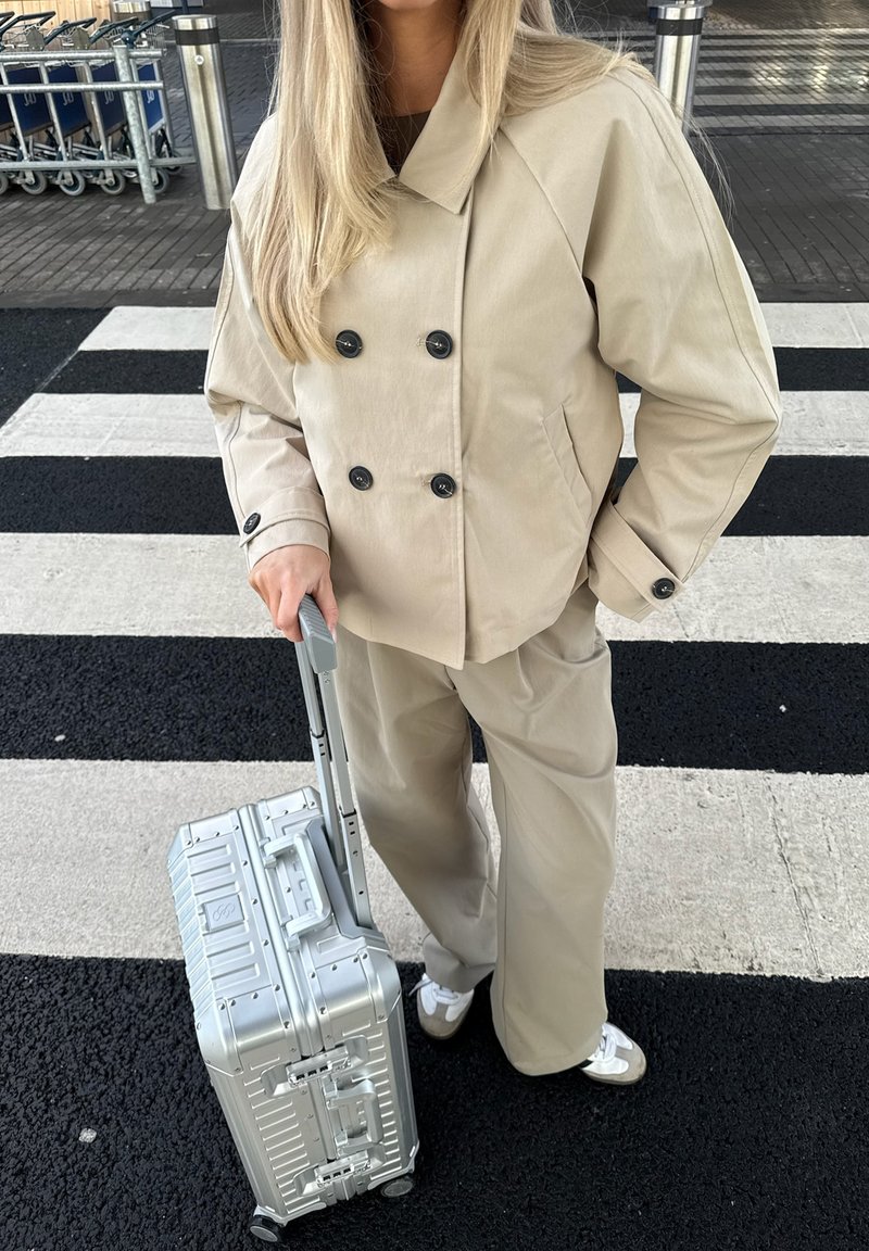 Person iført beige frakke og bukser, der holder en sølvfarvet kuffert på hjul på et sort-hvidt stribet fodgængerfelt nær bagagevogne.
