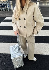 Beige kaksirivinen takki, yhdistelemällä housut, pitää hopeista matkalaukkuasi kuljetusväylällä, jossa on kontrastina musta-valkoinen pinta.