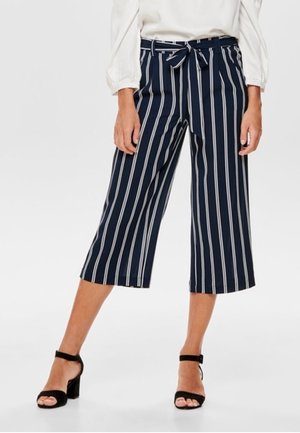 Femme portant un pantalon culotte bleu marine avec des rayures blanches verticales, une ceinture nouée à la taille, un chemisier blanc à manches longues et des talons noirs à bride de cheville.