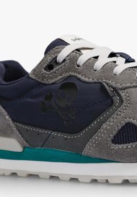 Zapatilla con parte superior de ante gris y textil azul marino. Presenta un logo de calavera y huesos cruzados en negro, cordones blancos y un acento en color aguamarina en la suela.