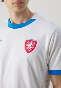 Balta sporta kreklā ar zilu apkakli un piedurknēm, kreisajā krūtī ir sarkans vairogveida logo, kas attēlo lauvas. Tekstūru audums.