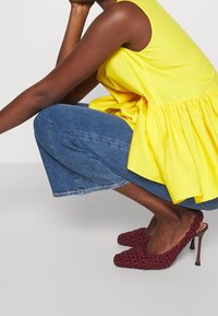 Mujer agachada, vistiendo una blusa sin mangas amarilla, jeans cortos azules y zapatos de tacón alto slingback de mimbre burdeos.