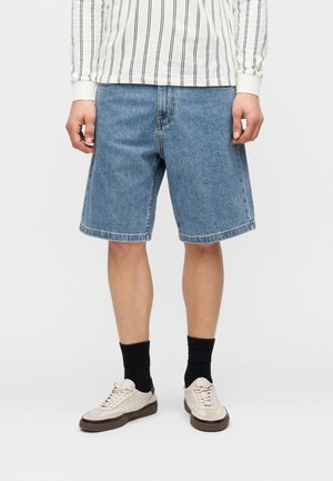 Hombre que lleva pantalones cortos de mezclilla azul hasta la rodilla, calcetines negros, zapatillas beige y una camisa blanca de manga larga con rayas negras verticales.