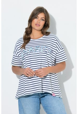 Vrouw met lang bruin haar die een witte en marineblauw gestreepte shirt met de tekst "SEASIDE" en lichtblauwe spijkerbroek draagt, staand tegen een witte muur.