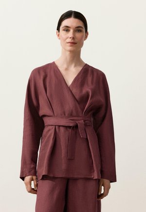 Kvinde med mørkt hår bundet tilbage iført en bordeauxfarvet, langærmet slå-om-bluse med matchende bælte og løstsiddende bukser mod en ensfarvet baggrund.