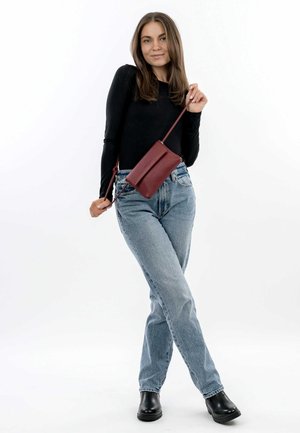 Bolso cruzado de cuero rojo con una textura suave, diseño minimalista, forma geométrica y correa ajustable, llevado con jeans de color azul claro y botas negras.