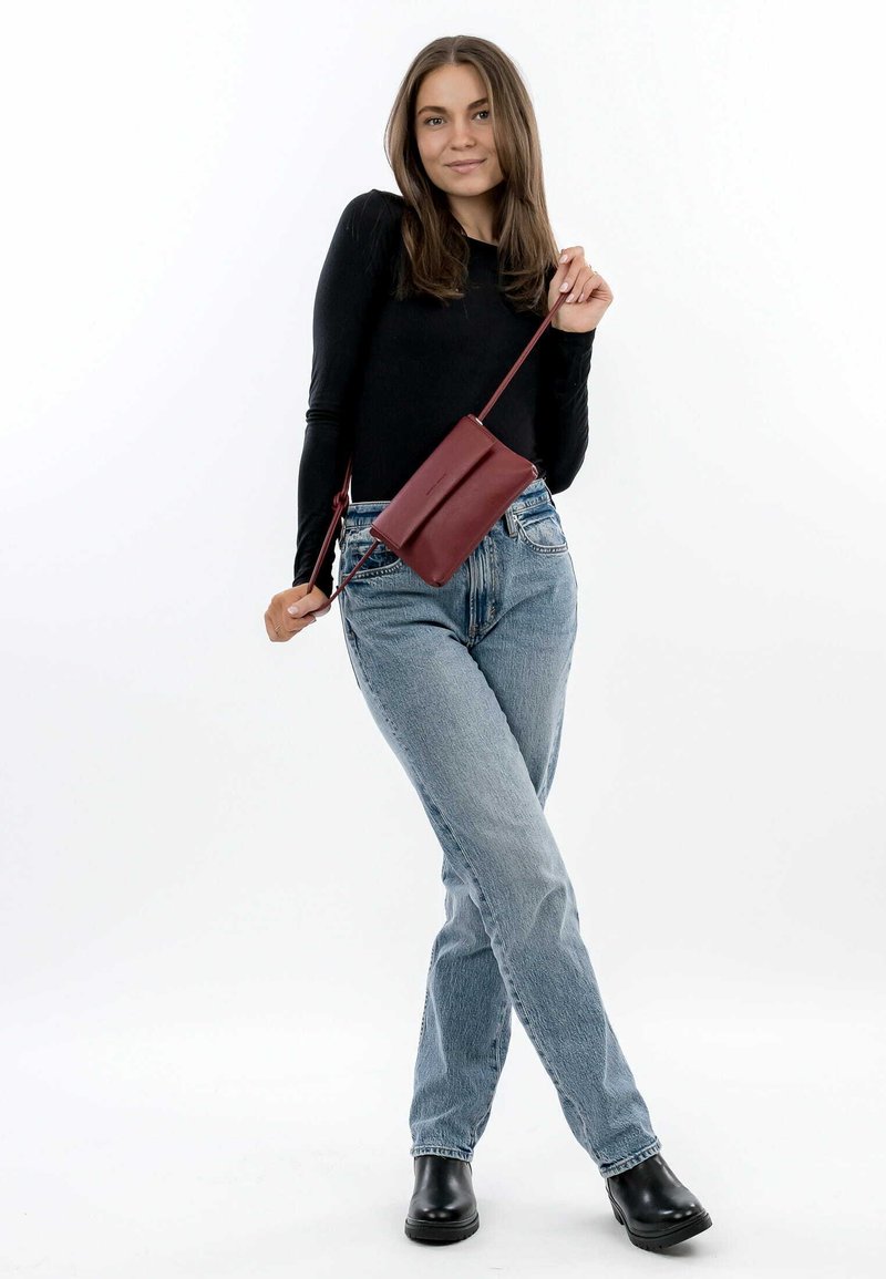 Red leather crossbody bag with a smooth texture, minimalist design, geometric shape, and adjustable strap, worn with light blue jeans and black boots.