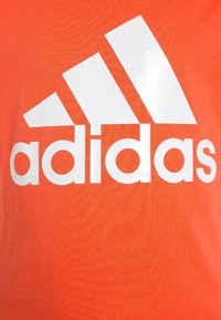 Pomarańczowy bawełniany T-shirt z dużym białym logo Adidas, zawierającym trzy poziome paski nad nazwą marki w pogrubionym tekście.
