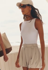 Femme portant un haut blanc sans manches, un short beige plissé avec une ceinture, une casquette beige et des lunettes de soleil, debout près d'une rambarde au bord de la plage.