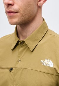 Κοντινό πλάνο ατόμου που φοράει καφέ πουκάμισο με κουμπιά, με τσέπη στο στήθος με φερμουάρ και λευκό λογότυπο The North Face στο στήθος.