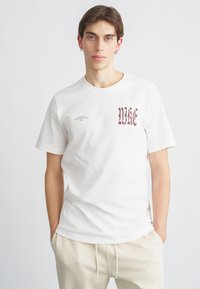 Biała bawełniana koszulka z logo Nike w kolorze bordowym, napisanym gotycką czcionką, z drobnym detalem tekstowym, okrągłym dekoltem i krótkimi rękawami.