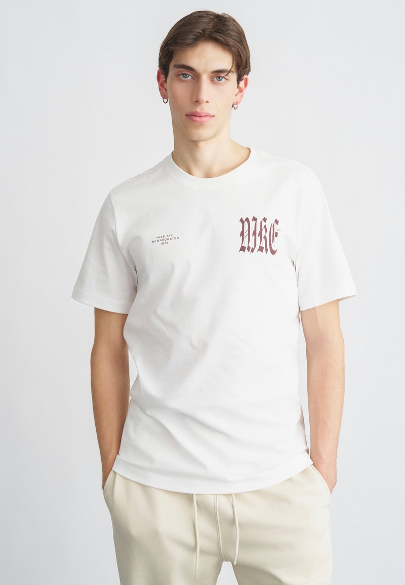 Biała bawełniana koszulka z logo Nike w kolorze bordowym, napisanym gotycką czcionką, z drobnym detalem tekstowym, okrągłym dekoltem i krótkimi rękawami.