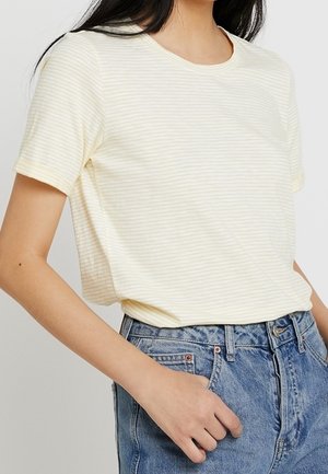 Femme portant un t-shirt à manches courtes rayé jaune clair et blanc, rentré dans un jean bleu taille haute, avec une main dans la poche.