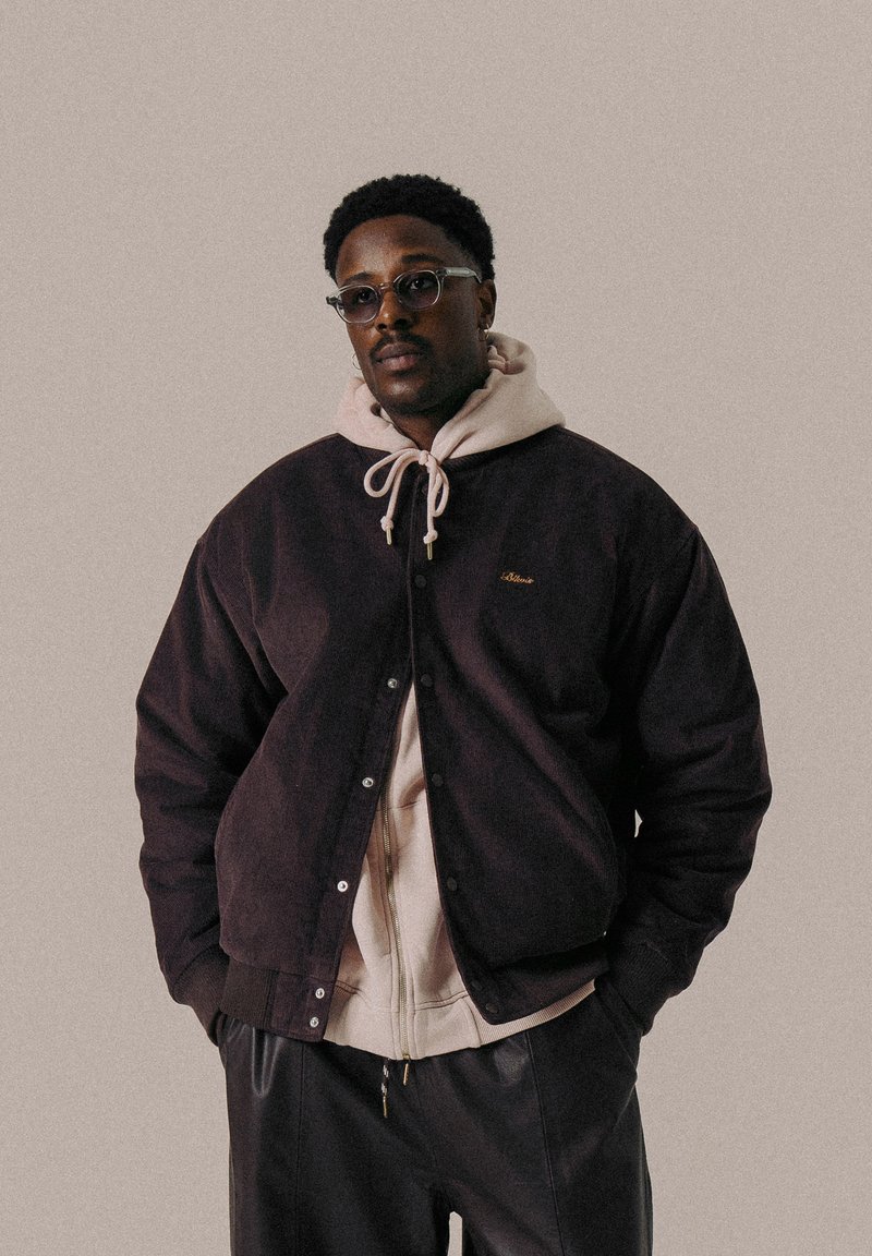 Dark purple corduroy jacket with snap buttons, layered over a light hoodie with a drawstring. Black pants complete the outfit.