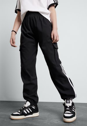Personne portant un pantalon de survêtement Adidas noir à bandes blanches et des baskets Adidas noires et blanches, debout devant un fond uni.