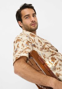 Camicia a manica corta in beige chiaro con motivi astratti marroni e blu, realizzata in tessuto leggero. Modello seduto su una sedia di legno.