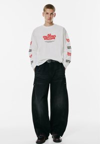 Junge Person in einem weißen Langarmshirt mit roten und schwarzen Schriftzügen, schwarzen weiten Hosen, schwarzem Gürtel und dunklen Schuhen, die mit den Händen in den Taschen dasteht.