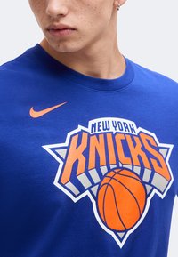 Νέος που φοράει μπλε T-shirt των New York Knicks με πορτοκαλί λογότυπο μπάσκετ και πορτοκαλί σήμα Nike.