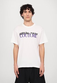 Biely bavlnený tričko s čiernym a fialovým textom "VERSACE JEANS COUTURE." Krátke rukávy, okrúhly výstrih, voľný strih.