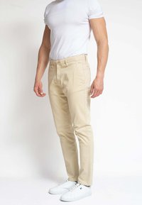 Hombre con pantalones beige ajustados, camiseta blanca y zapatillas blancas, de pie contra un fondo blanco liso.