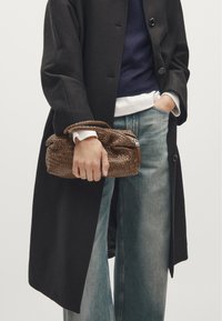 Gewebte braune Handtasche mit zwei Griffen, kompaktem rechteckigem Design und einem Reißverschluss. Getragen zu einem schwarzen Mantel und lockerer Jeans.