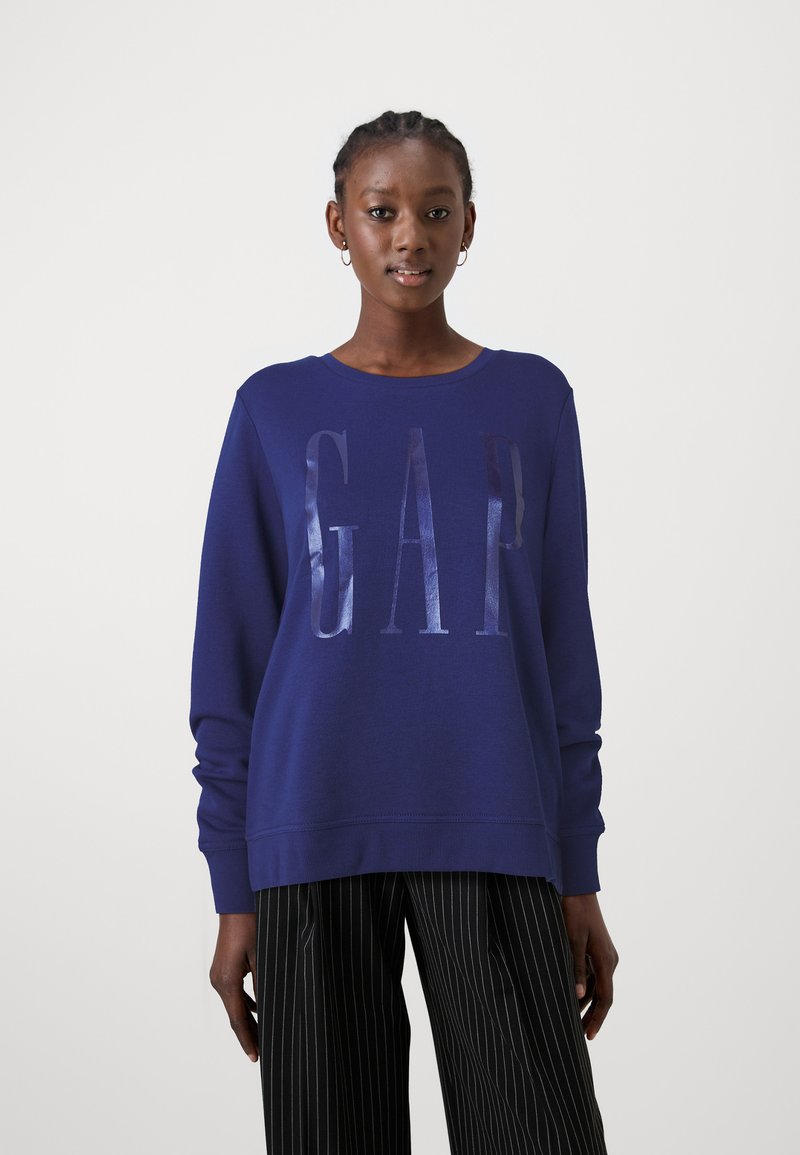 Tamsiai mėlynas džemperis su blizgiu "GAP" logotipu, apvalia iškirpte ir laisvu siluetu. Dėvimas su juodomis dryžuotomis kelnėmis.