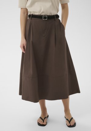 Osoba ubrana w brązową spódnicę midi z plisami, czarny pasek, beżowy top z krótkim rękawem i czarne sandały na cienkich paskach, stojąca na jasnym tle.