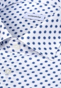 Wit overhemd met een kraag, voorzien van een blauw patroon van cirkelvormige stippen. Gestructureerde stof en knoopdetails zichtbaar nabij de kraag. Label aanwezig.