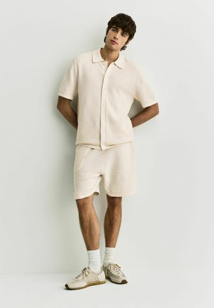 Ung mand står op ad en hvid væg iført en cremefarvet, tekstureret kortærmet skjorte med knapper, matchende shorts, hvide sokker og beige sneakers.
