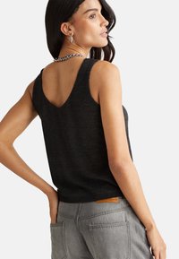 Top senza maniche nero con design a V sulla schiena, realizzato in materiale morbido. Abbinato a pantaloni grigi chiari con tasche posteriori evidenti e una etichetta in pelle.