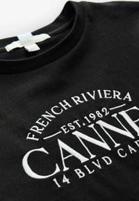 Tissu noir avec texte brodé en blanc : "FRENCH RIVIERA EST. 1982 CANNES 14 BLVD CAR".