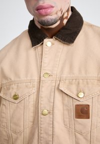 Chaqueta de lona beige con un cuello de pana marrón. Cuenta con cuatro bolsillos frontales, botones de hardware dorado y un parche de cuero con el logo.