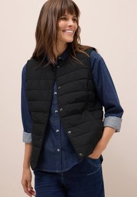 Gilet trapuntato nero con chiusure a pressione, indossato sopra una camicia in denim blu. Il gilet presenta un colletto alto e linee di cucitura orizzontali.