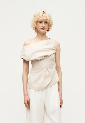 Άτομο με κοντά σγουρά ξανθά μαλλιά φοράει ασύμμετρο μπεζ τοπ και φαρδιά λευκά παντελόνια, στέκεται σε απλό φόντο.