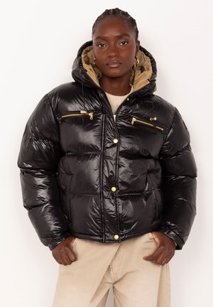 Femme portant une doudoune noire brillante avec des fermetures éclair et boutons dorés, une doublure de capuche beige et un pantalon beige sur fond blanc uni.