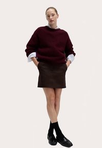 Pull oversize bordeaux à épaules tombantes, associé à une mini-jupe marron. Chaussettes noires et mocassins complètent le look. Textures lisses visibles.