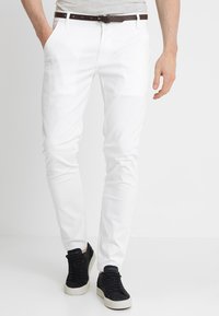 Pantalones de algodón blanco ajustados con una textura suave, detallados con trabillas para el cinturón y un cinturón marrón, combinados con zapatillas negras.