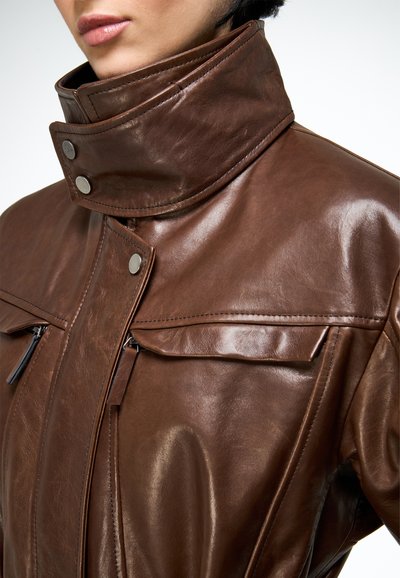 Gros plan d'une personne portant une veste en cuir marron avec un col montant fixé par des boutons-pression en métal et des poches poitrine zippées.