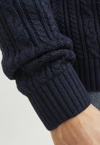 Navy blue knitted sweater sleeve showcasing a textured cable pattern and ribbed cuff detail, made from a thick, soft wool blend.