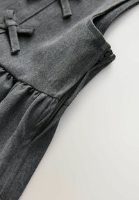 Tissu gris foncé avec une texture douce, présentant une fermeture éclair latérale et des détails froncés. De petits nœuds en tissu accentuent la partie supérieure.