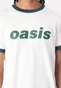 White cotton t-shirt with green ribbed collar and sleeves; features bold green text spelling "oasis" across the chest. Simple design.