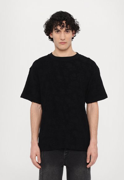 T-shirt noir à manches courtes avec un motif floral texturé, décolleté ras du cou et coupe décontractée, associé à un jean foncé.