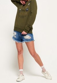 Sudadera verde oliva con texto y parches, combinada con pantalones cortos de mezclilla desgastados y zapatillas blancas con calcetines de red. Outfit casual.