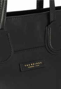 Borsa tote in pelle nera con texture liscia, doppi manici superiori e placca di branding dorata. Presenta un design semplice e strutturato con dettagli di cucitura.