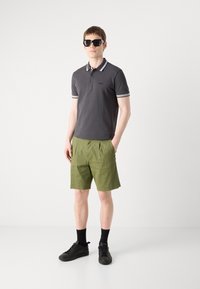 Tumši pelēks polo krekls ar baltu svītru apkakli, apvienots ar olīvzaļiem šortiem un melniem kedām. Modelis nēsā melnas saulesbrilles.