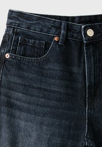 Gros plan sur un jean en denim bleu foncé avec poches avant, passants pour ceinture, rivets en cuivre et fermeture par bouton métallique à la taille.