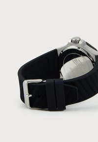 Bracelet de montre en caoutchouc noir avec un motif texturé, boucle en métal argenté et fond en acier inoxydable avec des détails gravés.
