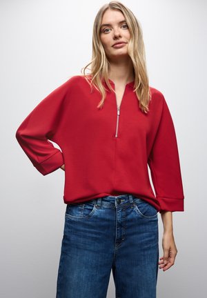 Roter Pullover mit halbhochgeschlossener Kragen, lockerer Passform und Dreiviertelärmeln, kombiniert mit blauen Denim-Jeans.