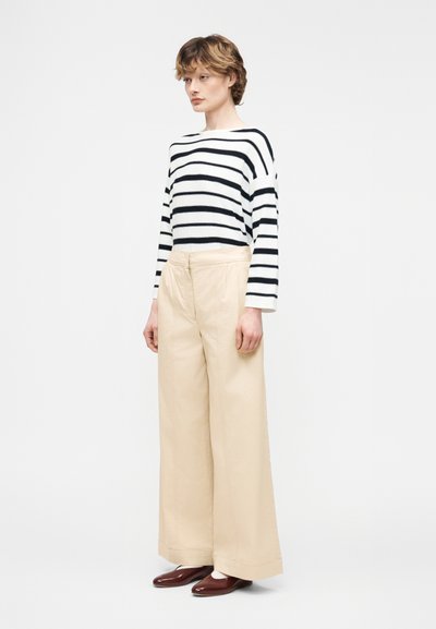 Άτομο που φοράει μαύρη και λευκή ριγέ μπλούζα με μακριά μανίκια, μπεζ παντελόνι φαρδύ στο κάτω μέρος και καφέ παπούτσια, στέκεται μπροστά σε μια απλή ανοιχτή φωτεινή επιφάνεια.